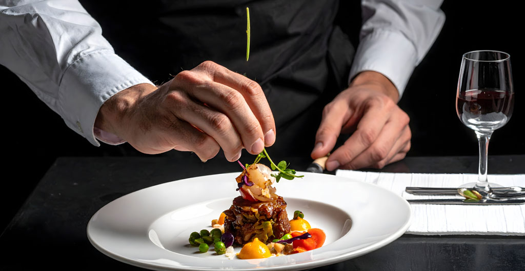 Chef cuisinier en tenue professionnelle dressant avec précision une assiette de Café de Paris dans une cuisine de brasserie moderne en inox, regard concentré sur le plat